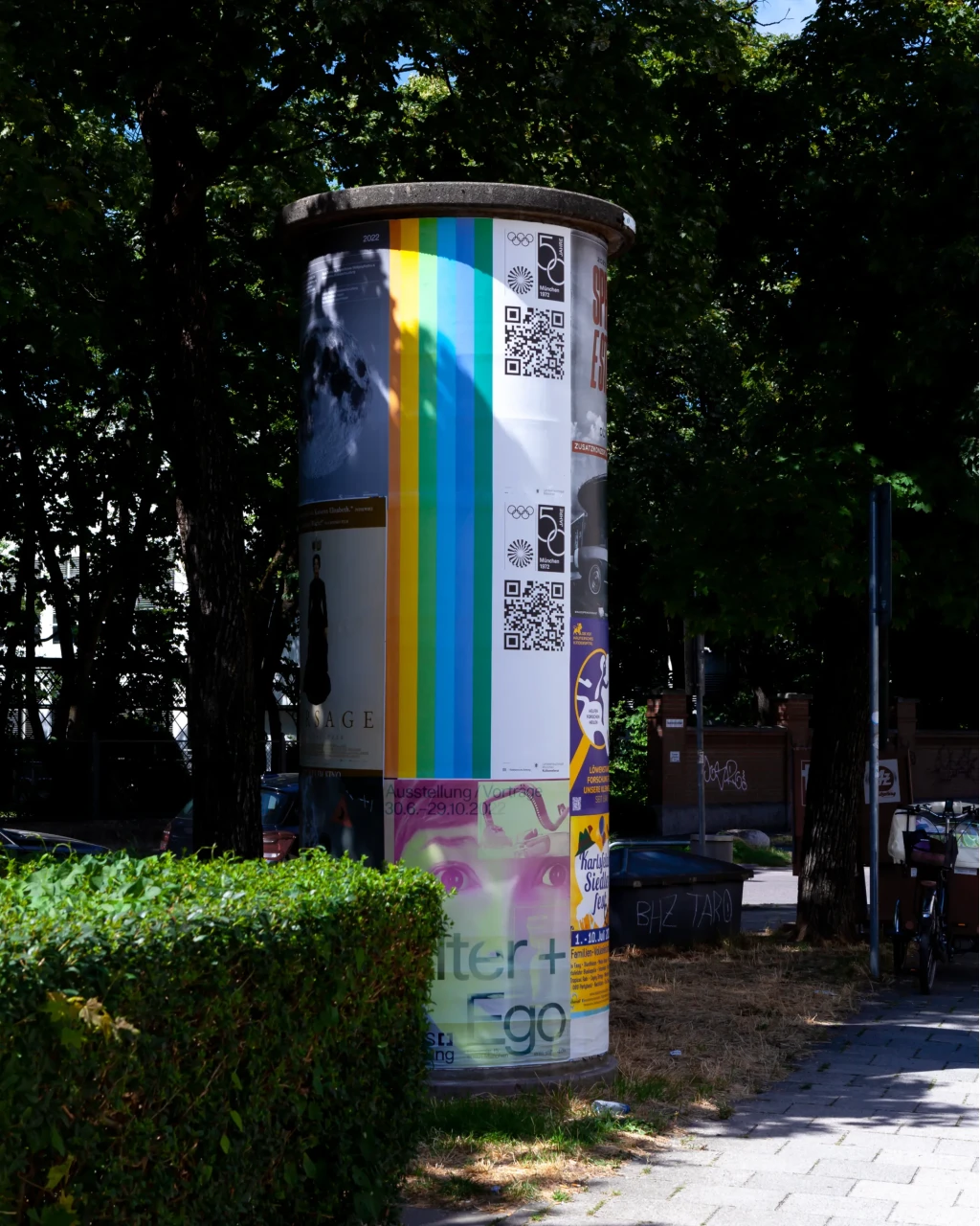 An einer Litfassäule kleben zwei Plakate des 50-jährigen Jubiläums der Olympischen Spiele in München mit bunten Streifen