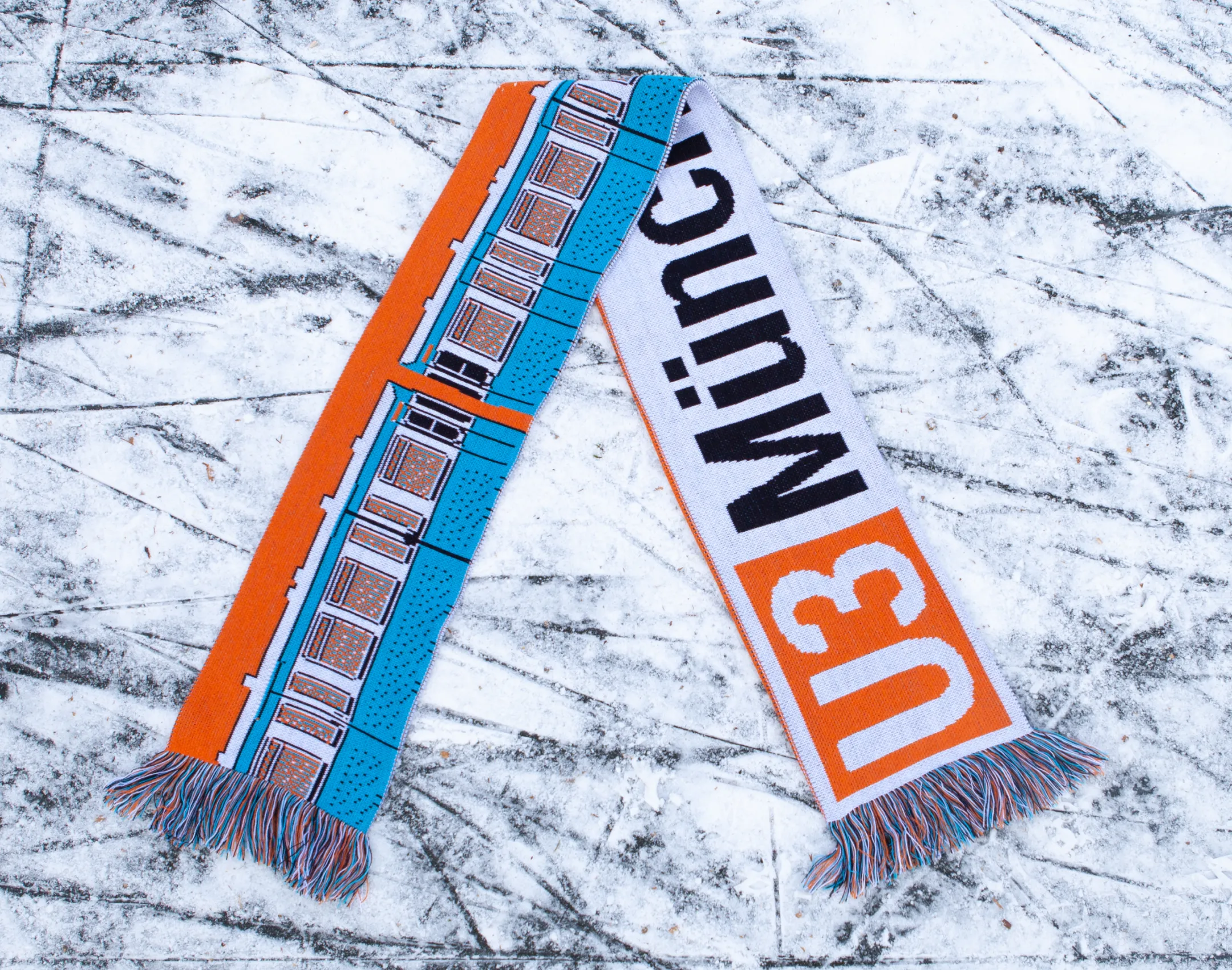 Orange-blauer Fußballlschal, der auf zugrfrorenem Kanal liegt. In U-Bahn Design der Schriftzug "U3 Münchener Freiheit"