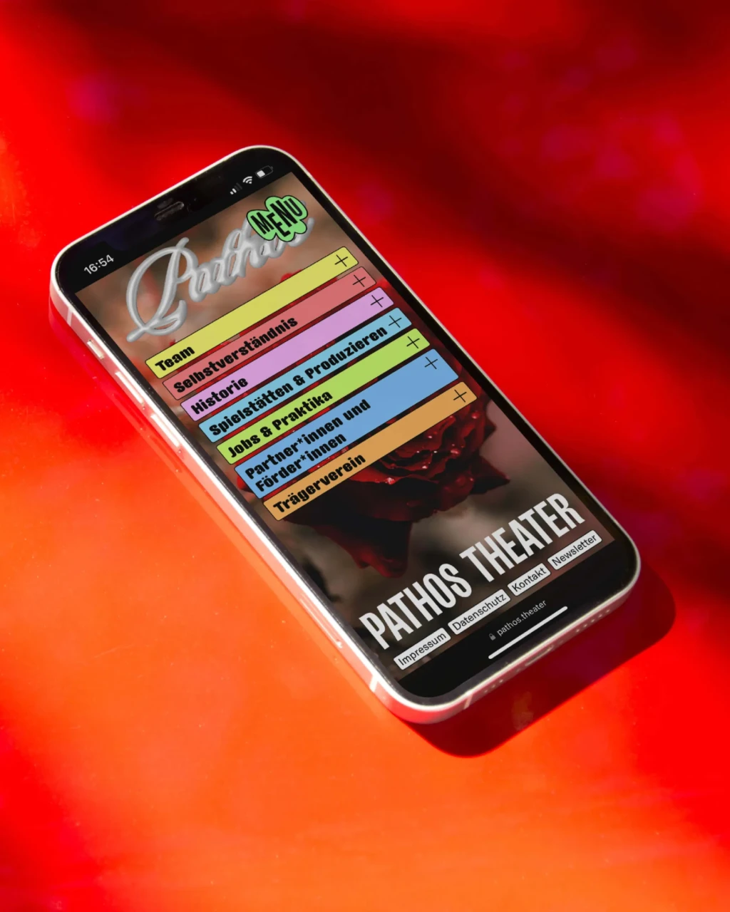 die Startseite von www.pathos.theater auf einem Smartphone, das auf einem roten Untergrund liegt