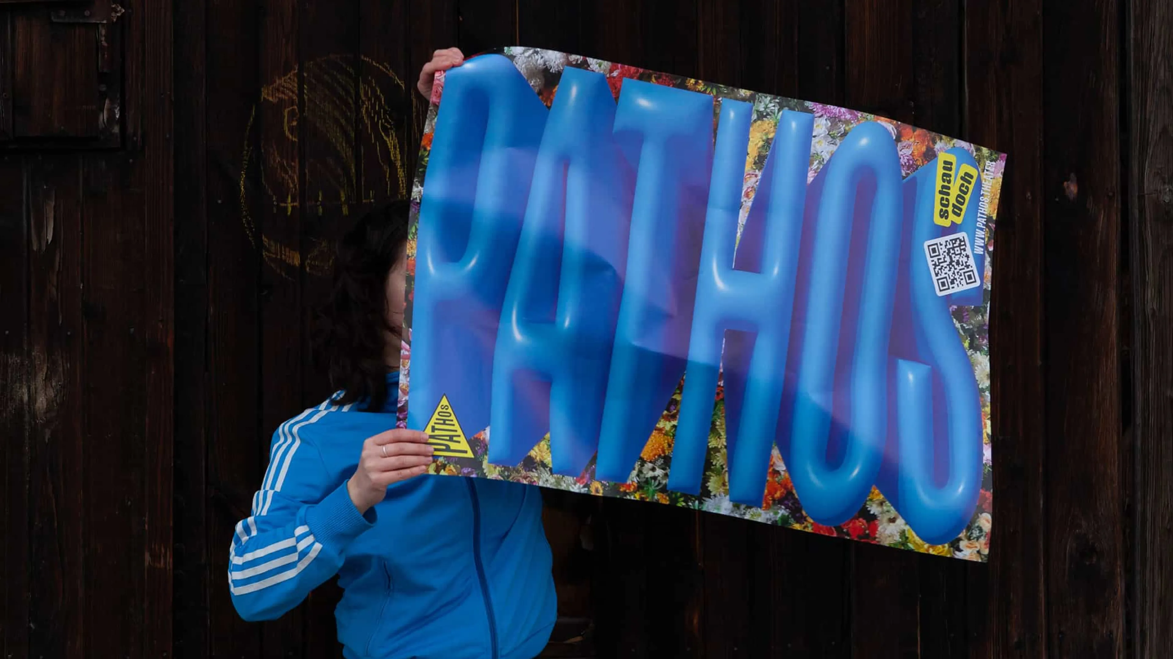 Person in blauer Trainingsjacke hält ein Pathos Theater Plakat mit blauer Bubble-Type Schriftzug vor floralem Hintergrund