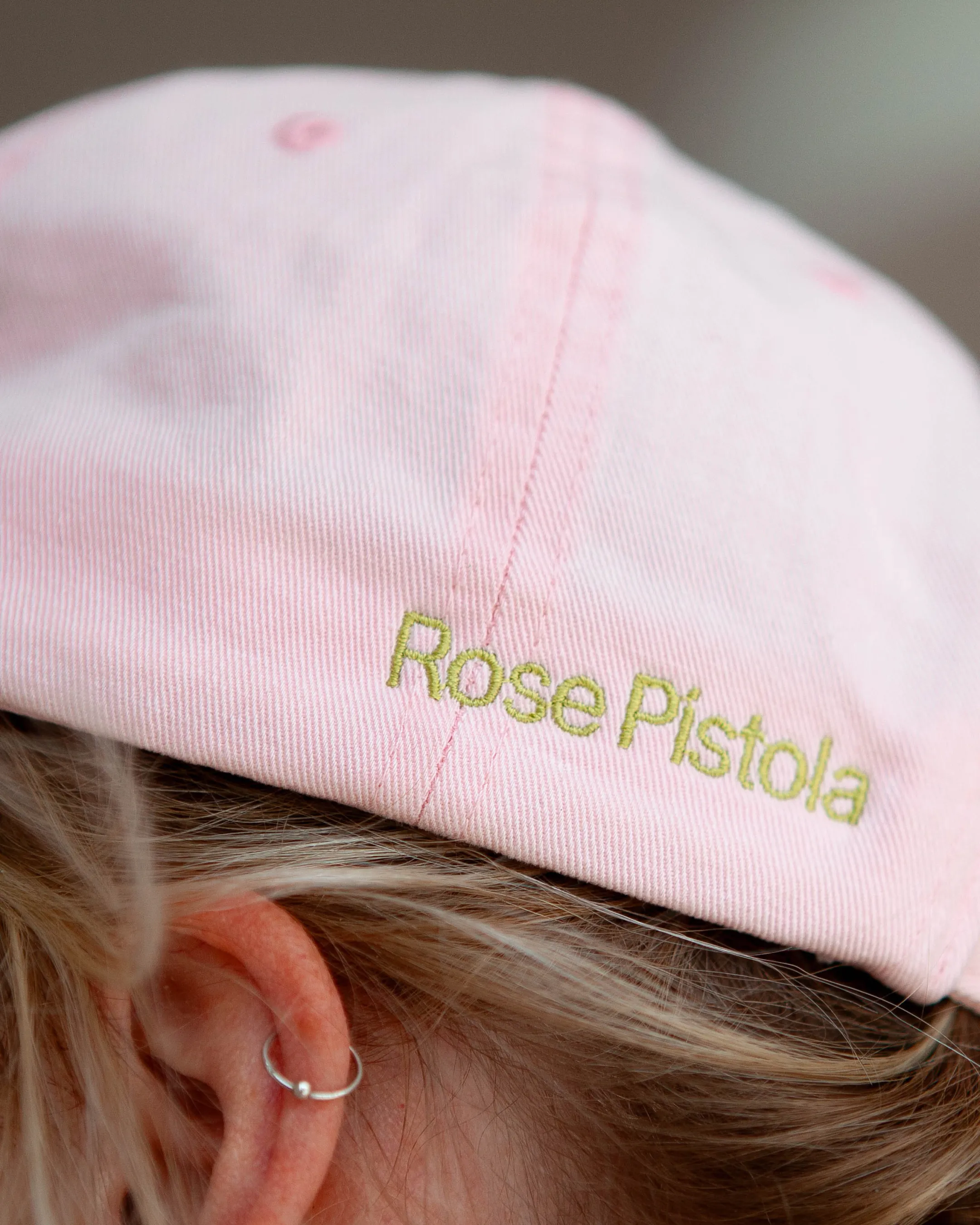 Detailaufnahme der rosa Merch Cap mit gold bestickter Rose Pistola Wortmarke auf dem Kopf einer Person