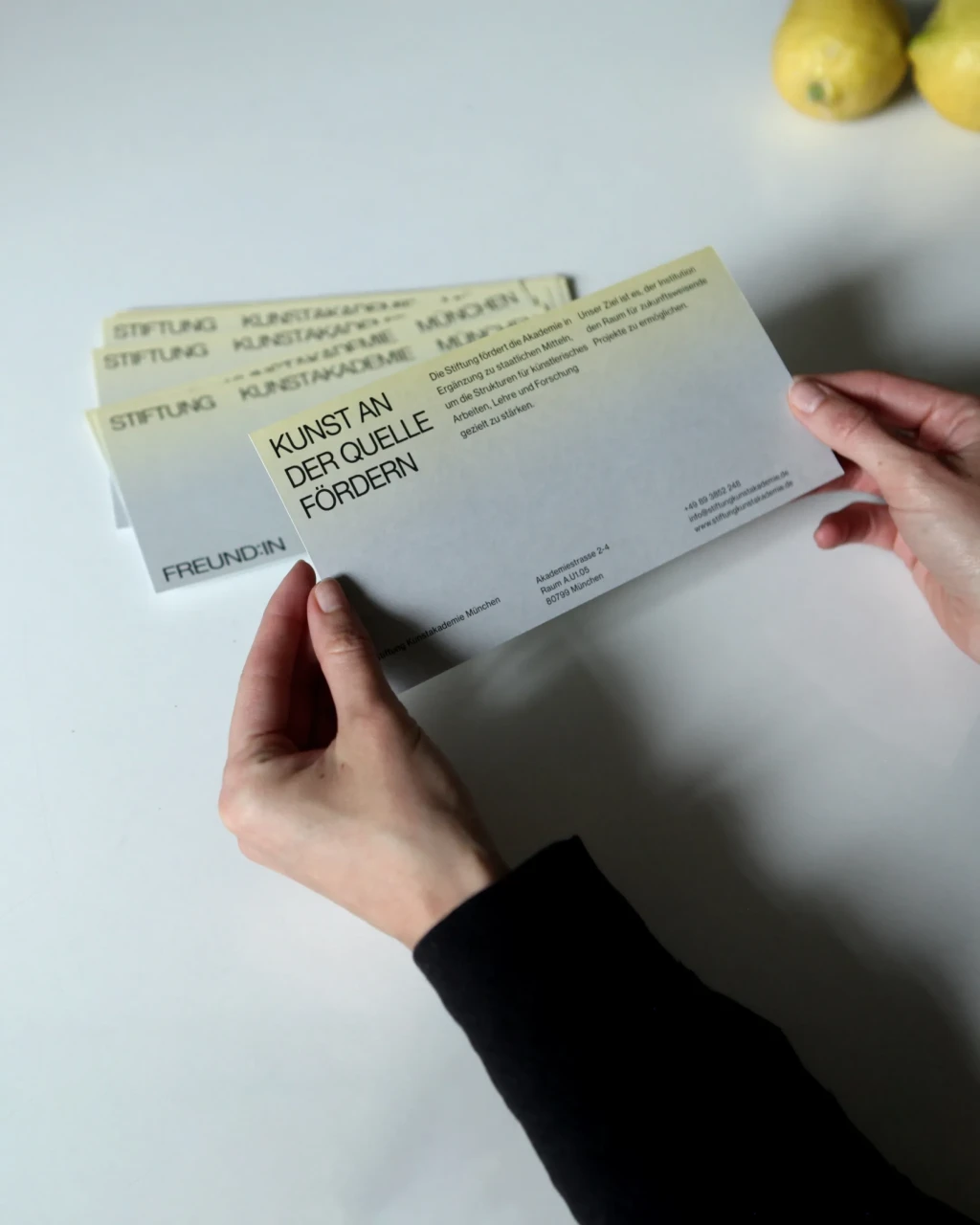 Infoflyer der Stiftung Kunstakademie München mit grau-gelbem Farbverlauf und Motto: Kunst an der Quelle fördern